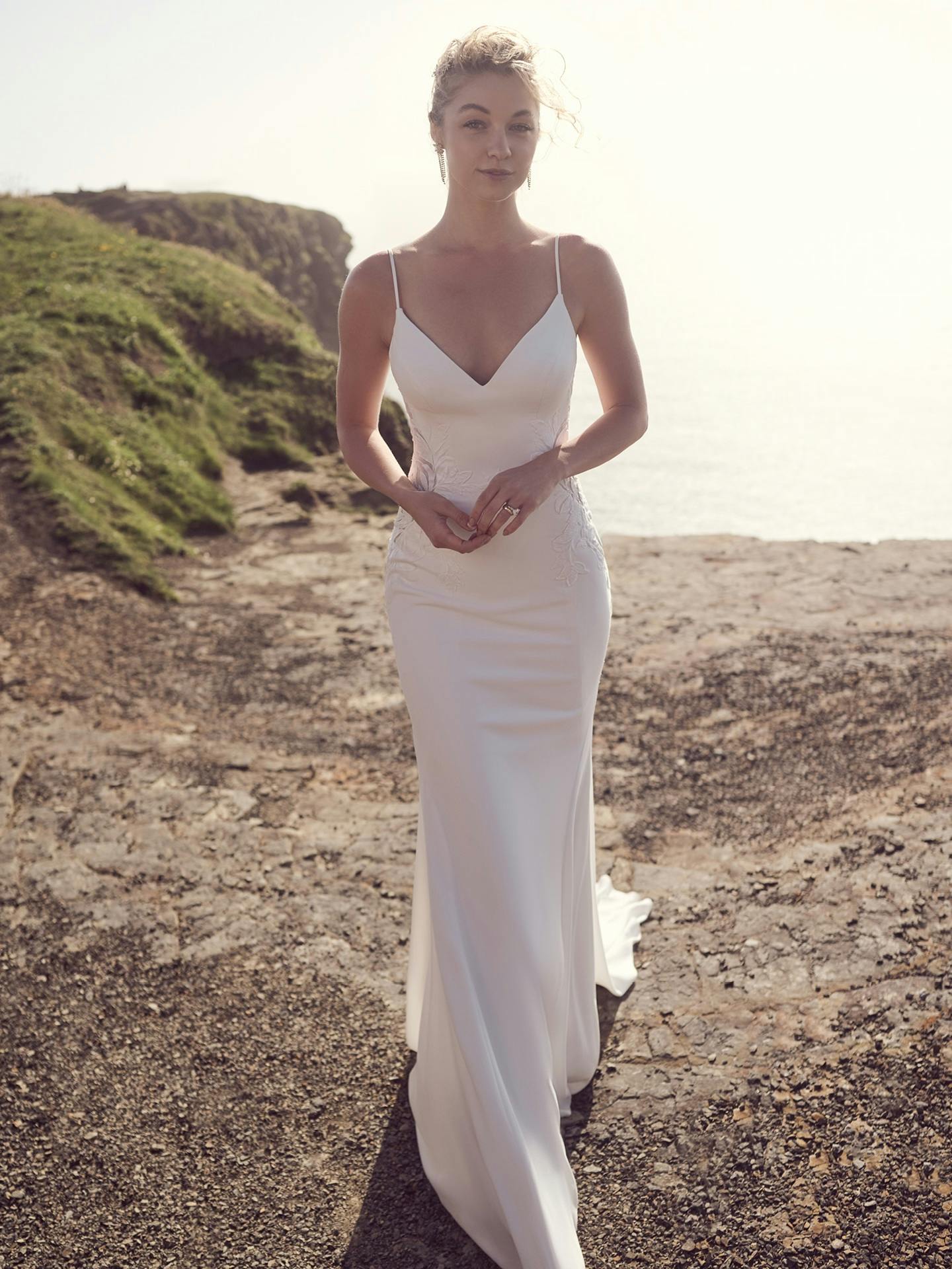 Woman in a white wedding dress standing on a rocky path with greenery and a clear sky in the background.