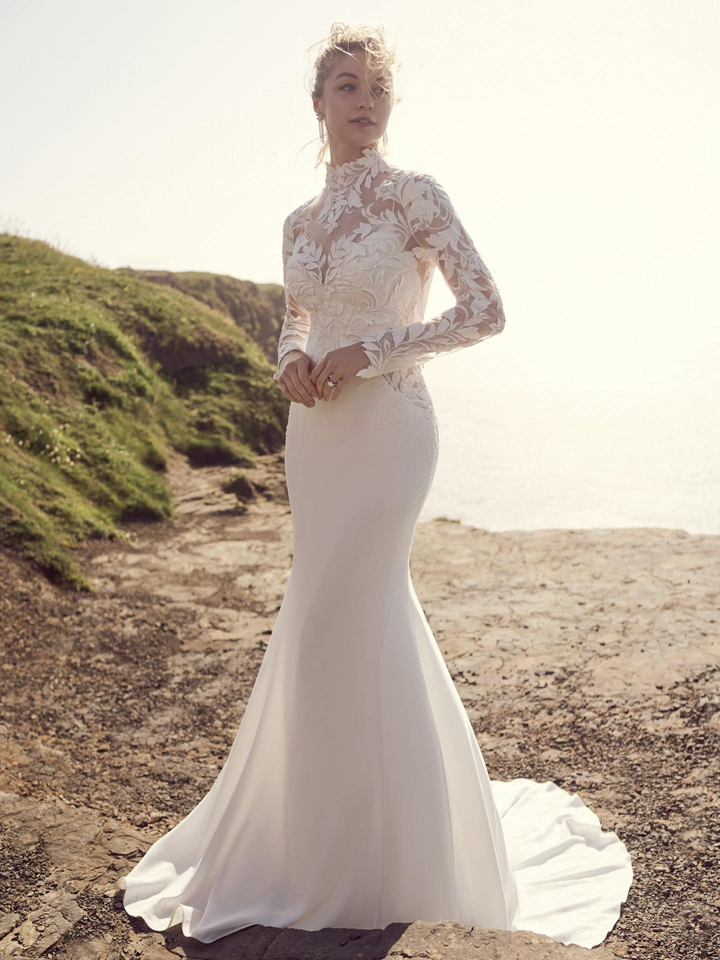 Woman in a white lace wedding dress standing on a beach.