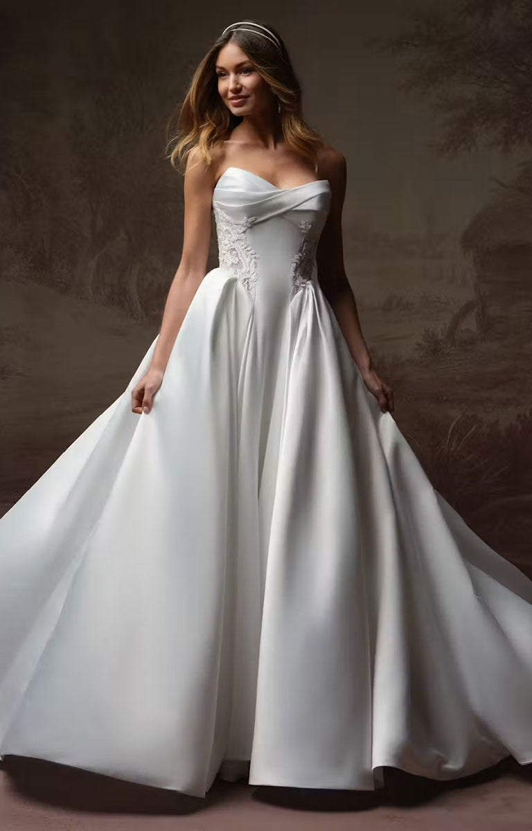 A woman modeling Gigi by Rebecca Ingram - 3D Floral Satin Ball Gown with Deep Basque Waist and Detachable Panniers with a fitted lace bodice and a full satin skirt, posed against a dark brown painterly backdrop.
