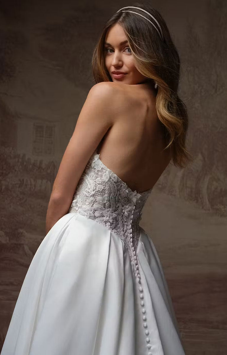 A woman wearing Gigi by Rebecca Ingram - 3D Floral Satin Ball Gown with Deep Basque Waist and Detachable Panniers with a sheer lace corset bodice and a long pearl-like down the back, photographed against a soft brown studio backdrop.