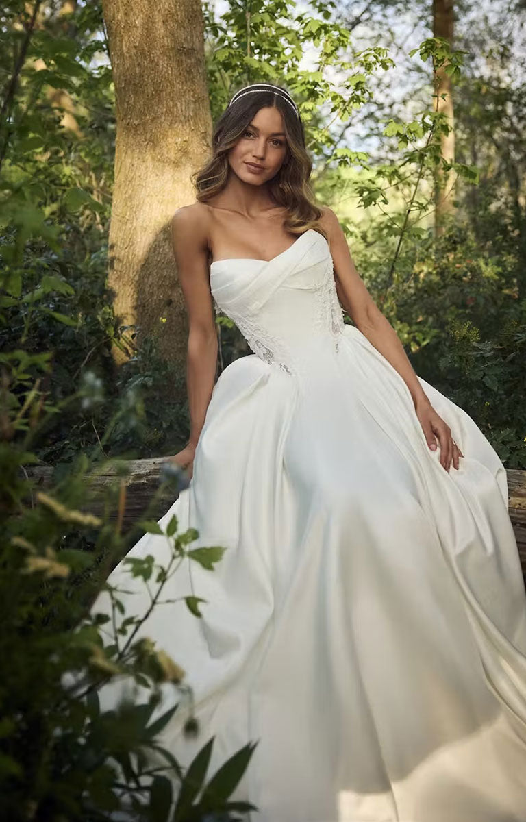 A woman wearing Gigi by Rebecca Ingram - 3D Floral Satin Ball Gown with Deep Basque Waist and Detachable Panniers with a fitted bodice and full ball gown skirt, posed outdoors in a sunlit forest setting.