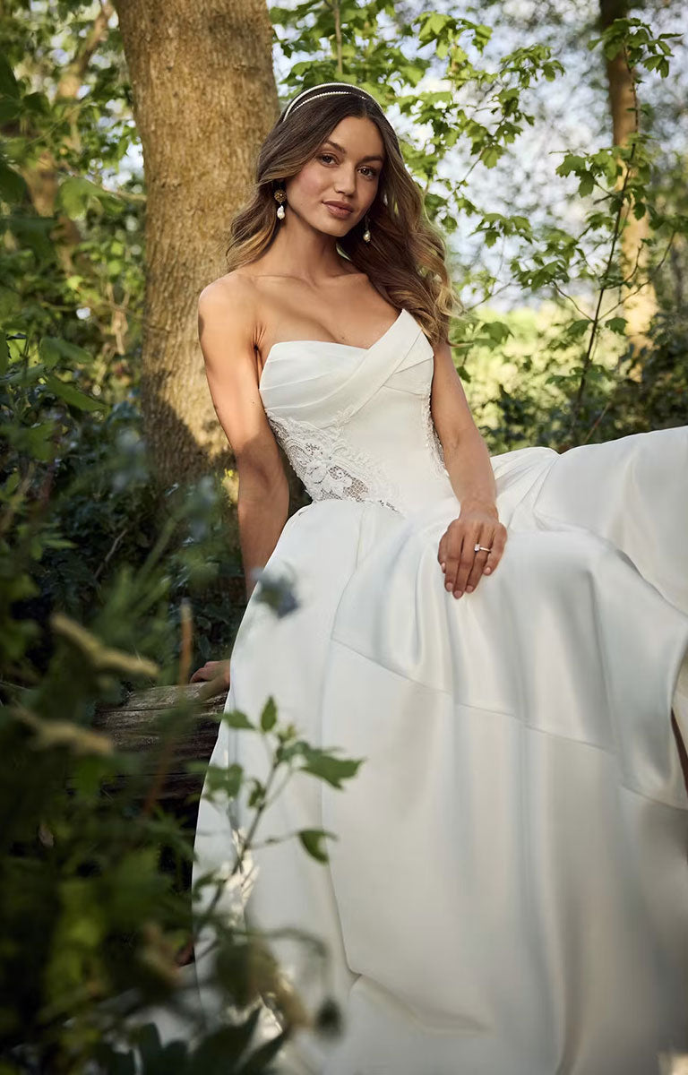 A woman wearing Gigi by Rebecca Ingram - 3D Floral Satin Ball Gown with Deep Basque Waist and Detachable Panniers with a fitted lace bodice and full satin skirt, posed outdoors among trees and greenery.