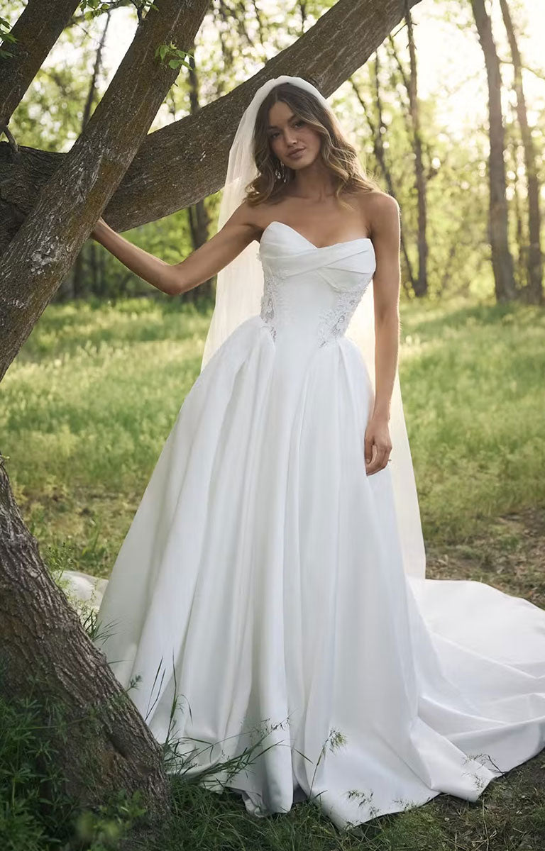 A woman wearing Gigi by Rebecca Ingram - 3D Floral Satin Ball Gown with Deep Basque Waist and Detachable Panniers with a sweetheart neckline, lace-embellished bodice, and full skirt, standing outdoors beside a tree in a sunlit grassy forest setting.