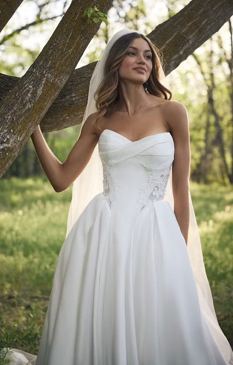 A woman wearing Gigi by Rebecca Ingram - 3D Floral Satin Ball Gown with Deep Basque Waist and Detachable Panniers with a sweetheart neckline, lace-embellished bodice, and full skirt, standing outdoors beside a tree trunk in a sunlit grassy area.