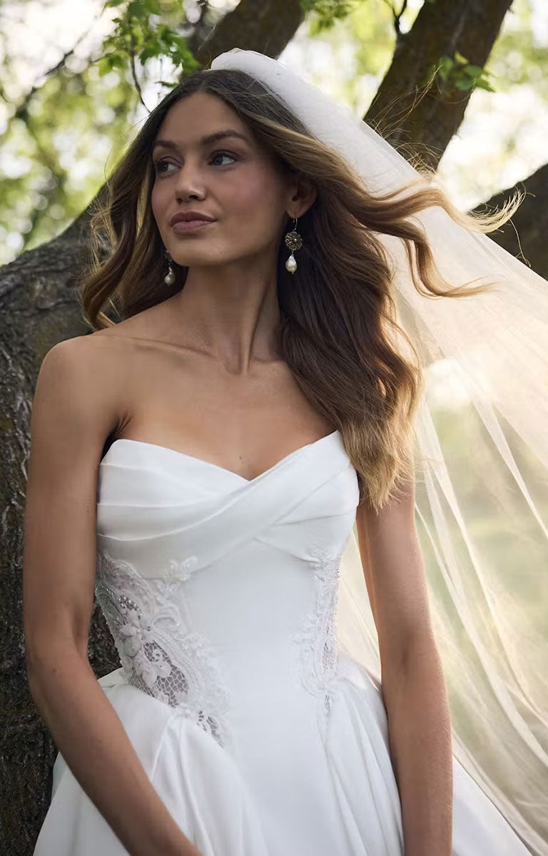 A woman wearing Gigi by Rebecca Ingram - 3D Floral Satin Ball Gown with Deep Basque Waist and Detachable Panniers with lace detailing and a long sheer veil, standing outdoors against a tree trunk with sunlight filtering through the background.