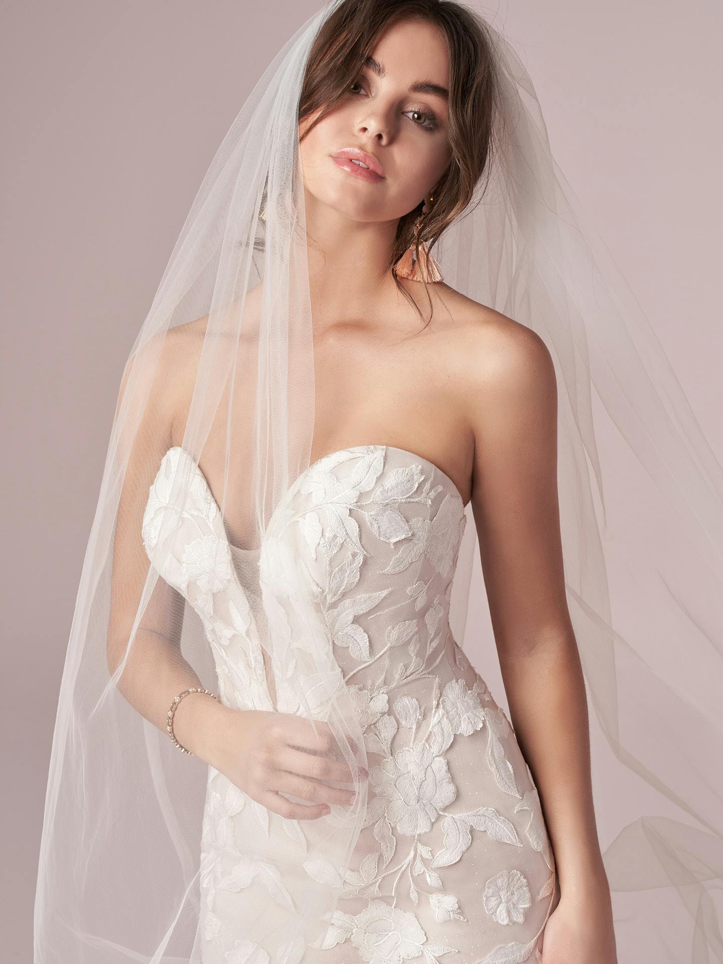 Woman wearing Hattie by Rebecca Ingram - a strapless wedding dress with floral details and a veil against a plain background