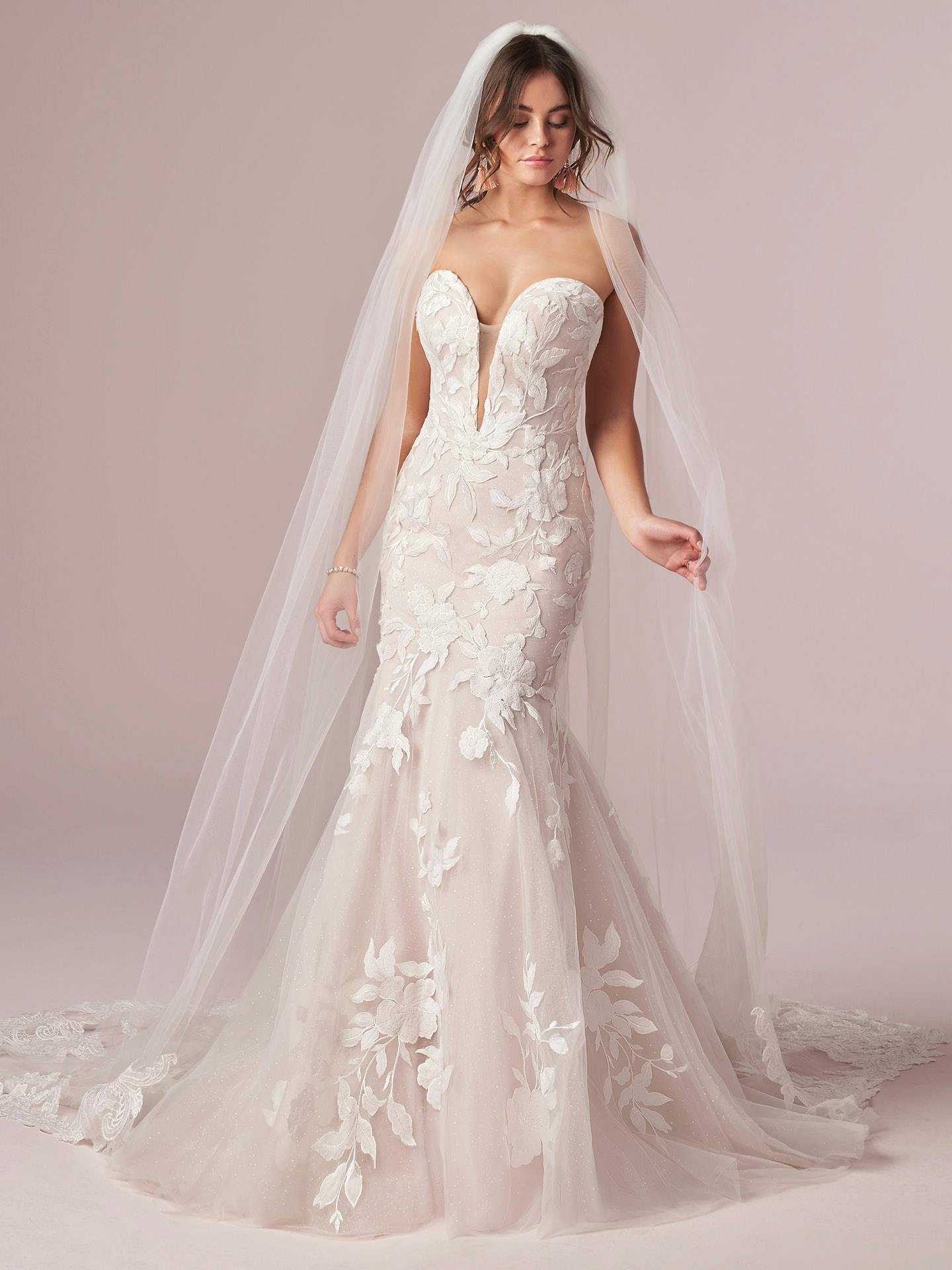 Woman wearing Hattie by Rebecca Ingram - a white wedding dress with floral details and a long veil on a plain background