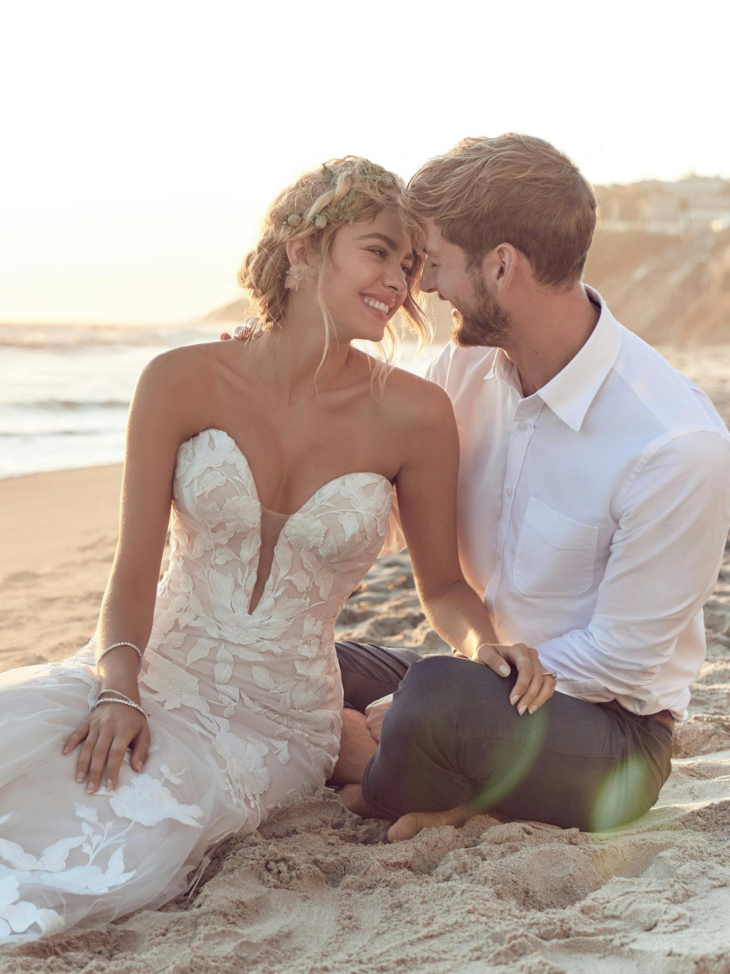 Couple sitting on a beach, woman in Hattie by Rebecca Ingram - a wedding dress, man in a white shirt.