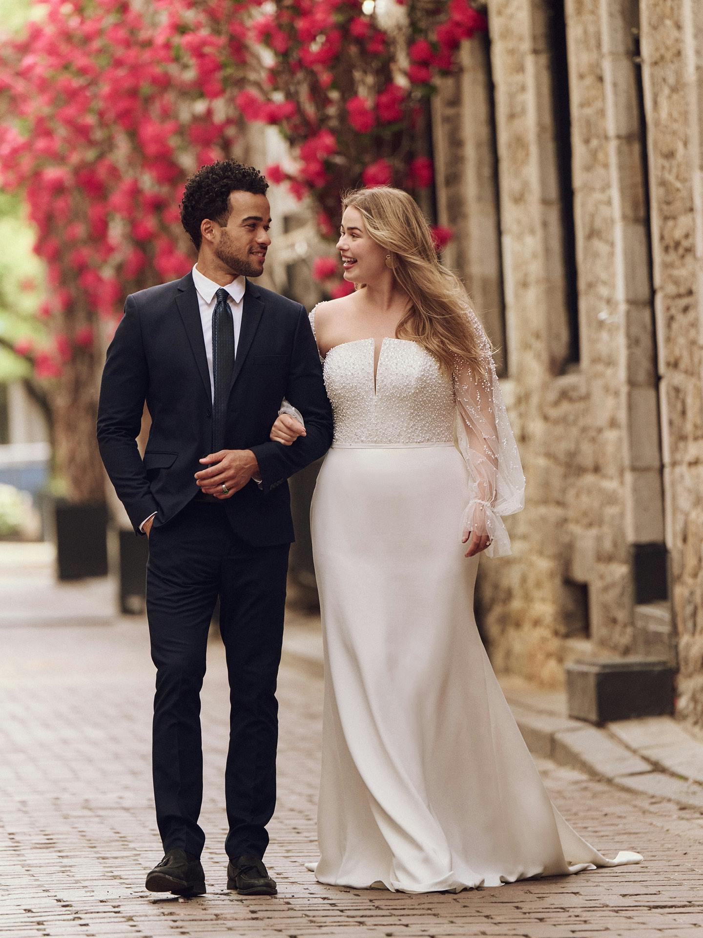 Man in a suit and woman in a white wedding dress walking together outdoors with flowers and stone building in the background.
