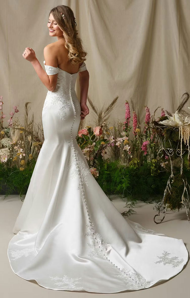 Woman in Miley by Rebecca Ingram - Off-the-Shoulder Fit-and-Flare Wedding Dress with Sequined Lace Accents standing in a garden setting with flowers and a cart.