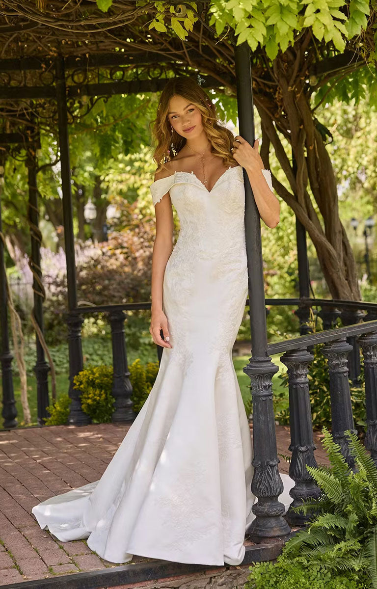 Woman in Miley by Rebecca Ingram - Off-the-Shoulder Fit-and-Flare Wedding Dress with Sequined Lace Accents standing outdoors under a pergola.