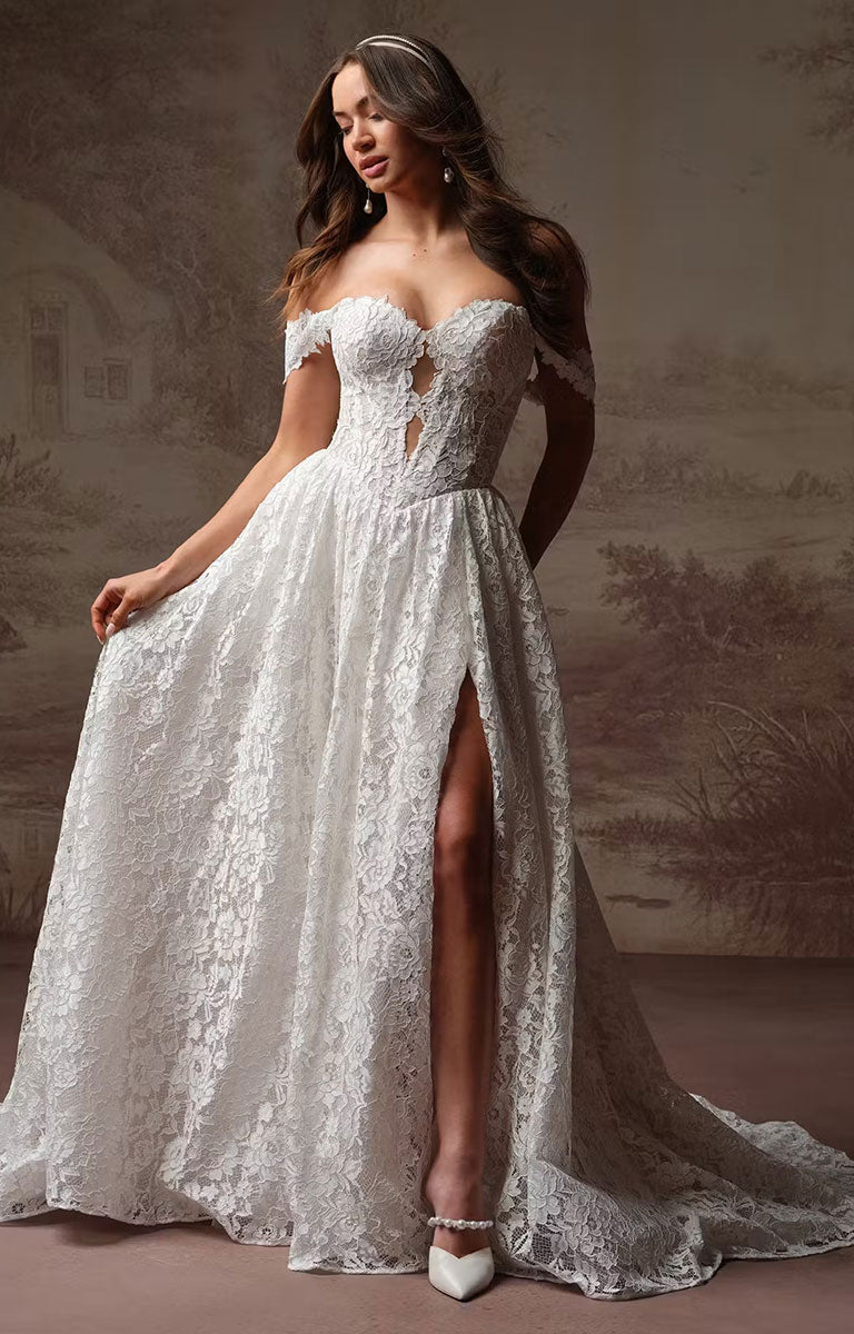 A woman models an Narelle by Rebecca Ingram - Plunging Lace Fit-and-Flare Wedding Dress with Side Slit with a sweetheart neckline, fitted bodice, and a high thigh slit, posed against a soft beige scenic backdrop.