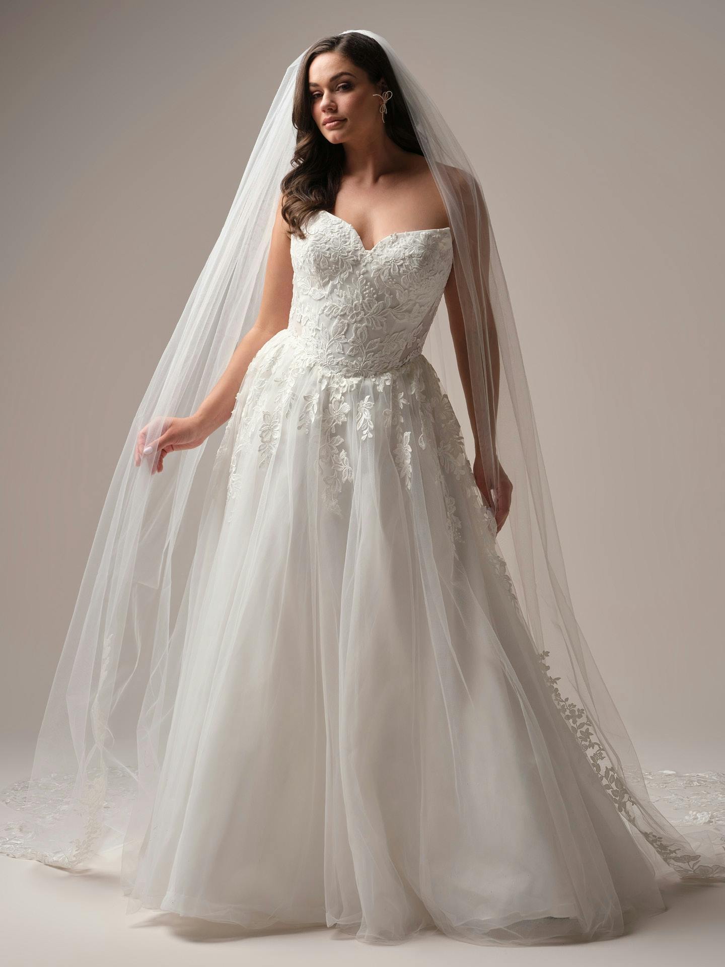 Woman wearing Paige by Rebecca Ingram -  a white wedding dress with a long veil against a neutral background