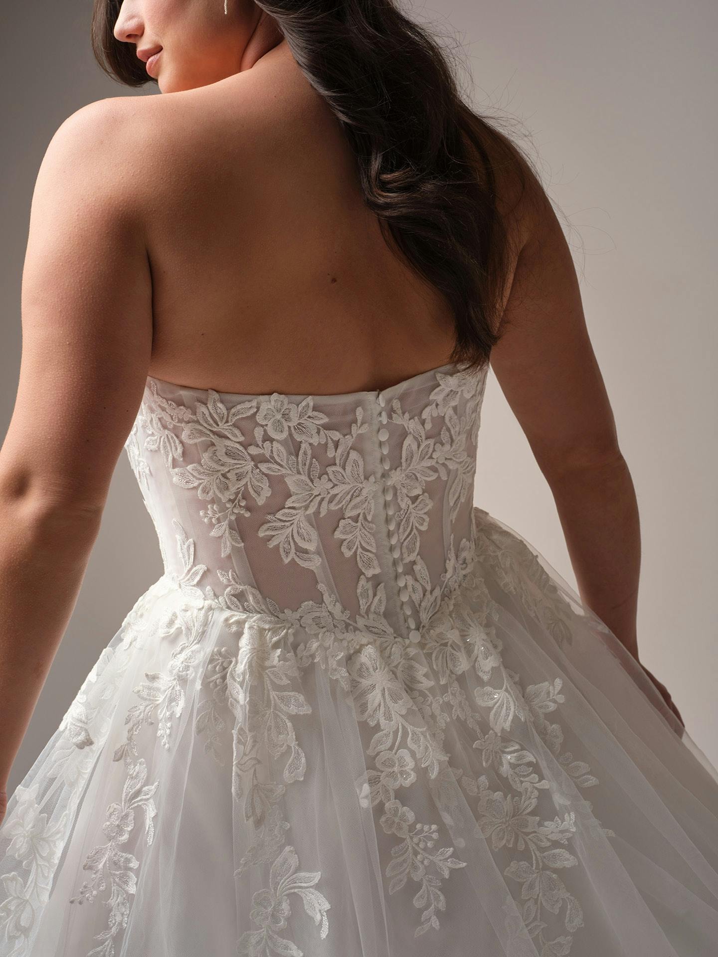 Close-up of a woman wearing Paige by Rebecca Ingram - a white lace wedding dress with a plain background