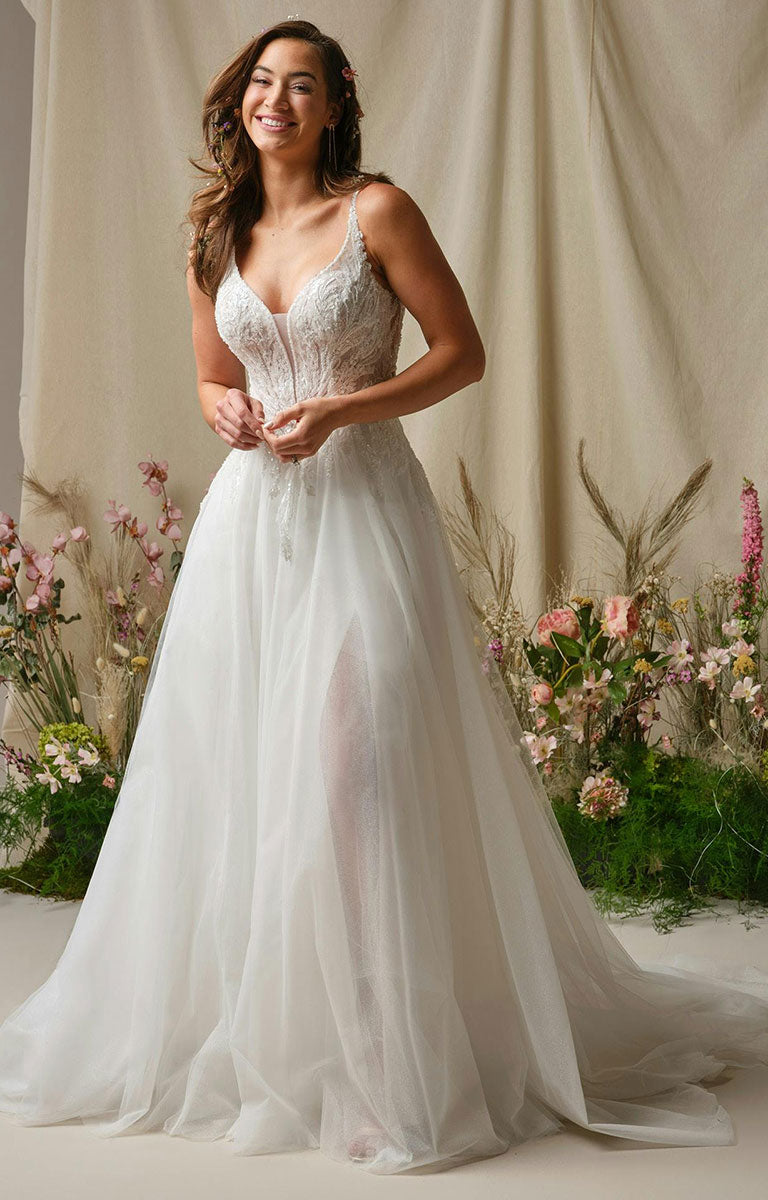 Woman in Valarie by Rebecca Ingram - Sparkle Lace A-Line Wedding Dress with Plunging V-Neck and Side Slit standing in front of floral decorations.