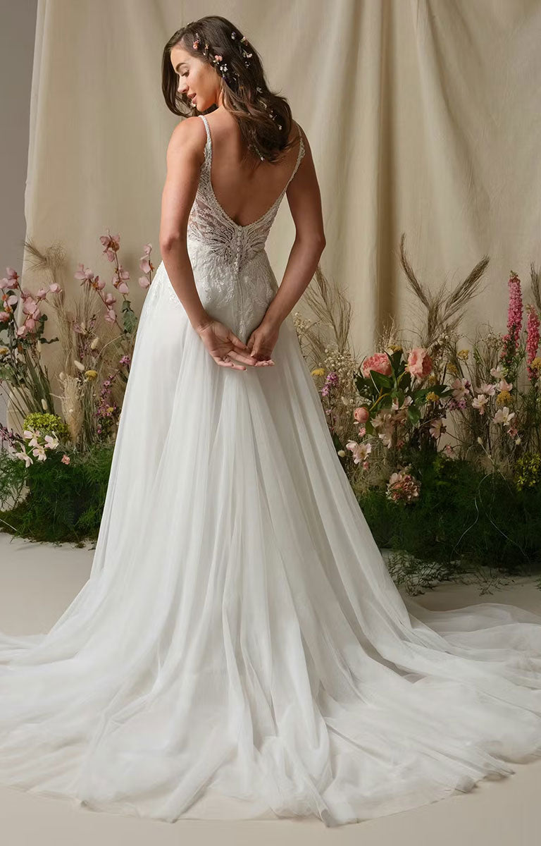 Woman in Valarie by Rebecca Ingram - Sparkle Lace A-Line Wedding Dress with Plunging V-Neck and Side Slit standing in front of floral decorations.