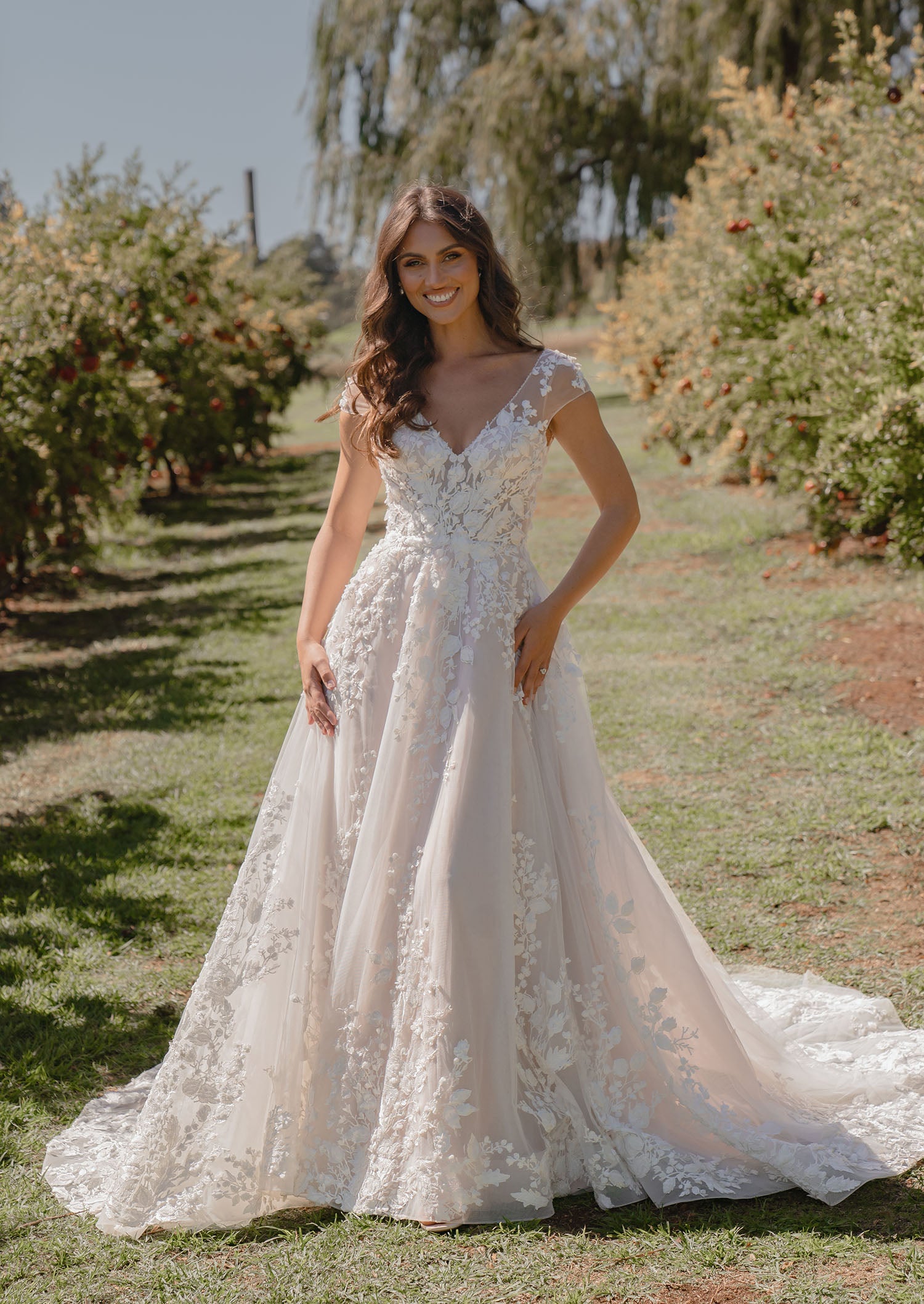 Woman in Sarah by Madi Lane, a lace wedding dress standing in an orchard.