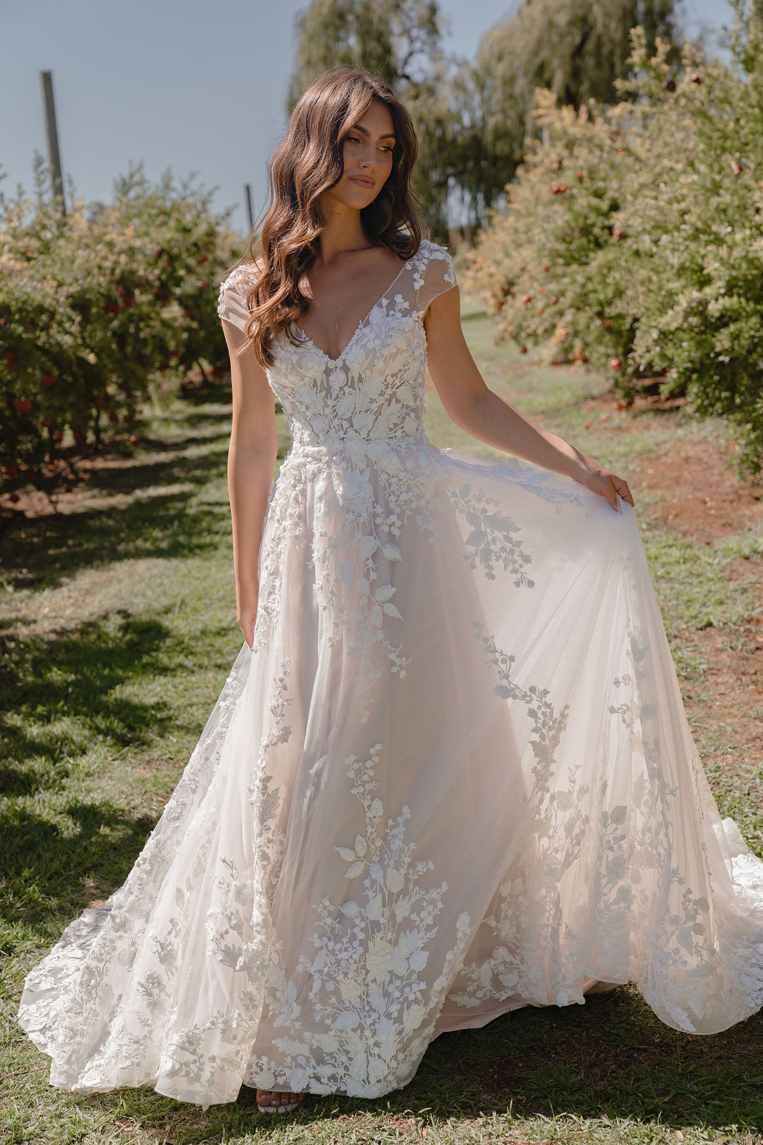 Woman in Sarah by Madi Lane, a white lace wedding dress standing in an outdoor setting with trees and grass.
