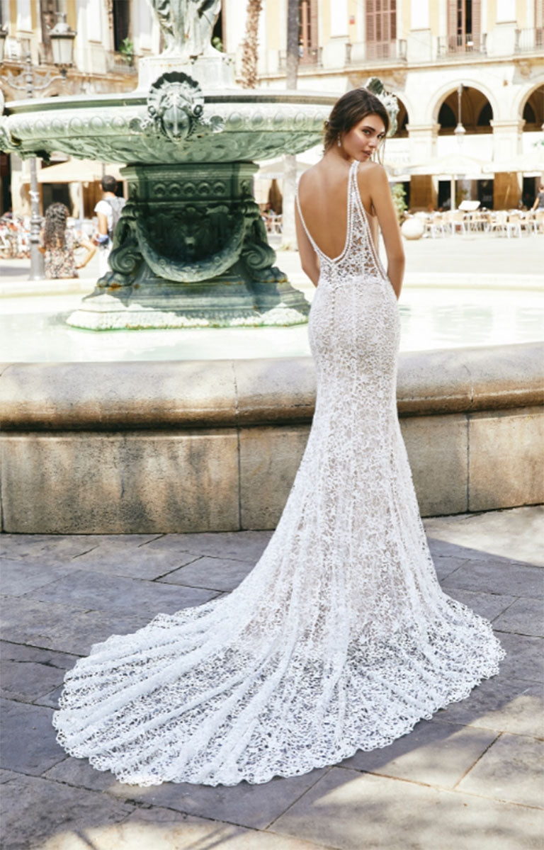 Woman standing in front of a fountain facing forwards and showcasing the rear of Sevyn by Veni Infantino - an elegant Fishtail Lace Wedding Dress with Illusion Bodice and Cathedral Train