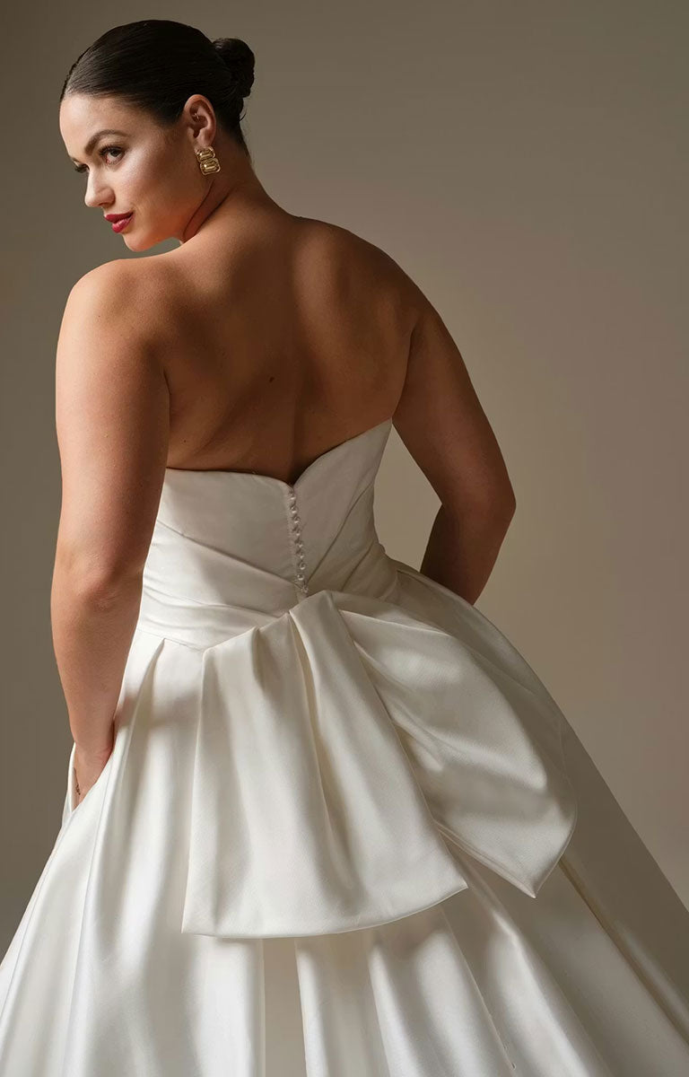 Woman wearing Kincaid by Sottero and Midgley - Ruched Satin Ballgown Wedding Dress with Pearl Details and Cat-Eye Neckline with a large bow detail on a plain background