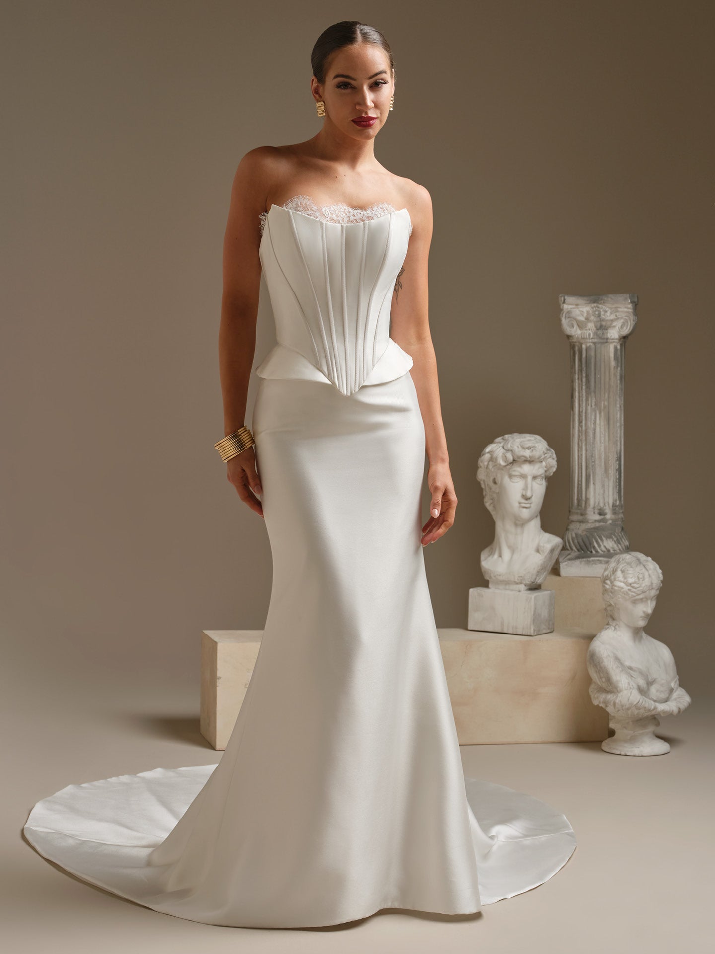 Woman in Lassen by Sottero and Midgley - Strapless Chantilly Lace Sheath Wedding Dress with Cat-Eye Neckline and Corset Bodice standing in front of classical sculptures.