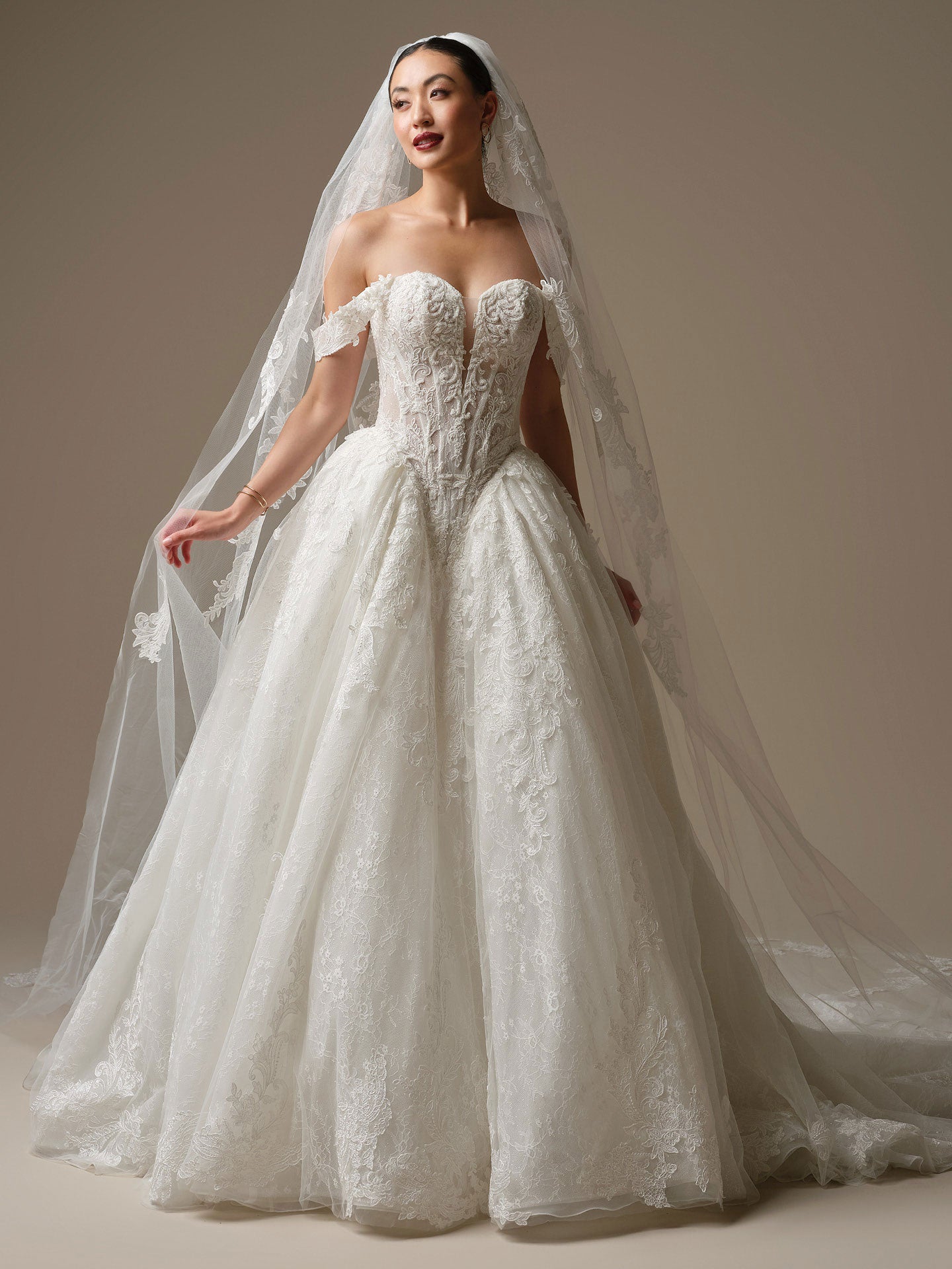 Woman wearing a white lace wedding dress with a long veil against a neutral background