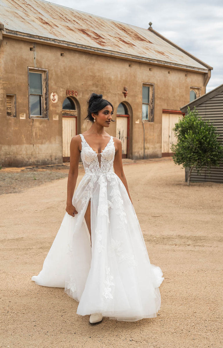 Woman in Tala by Madi Lane, a white wedding dress standing in front of an old building with a dirt ground.
