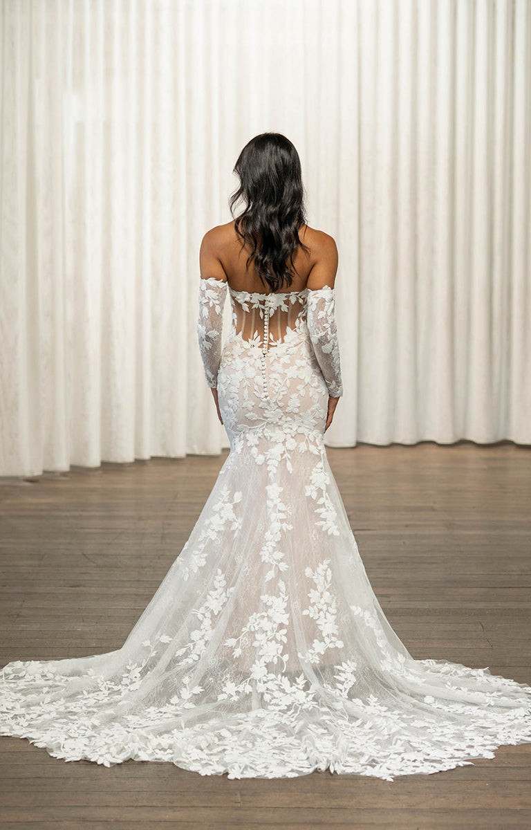 Woman wearing Thane by Madi Lane, a white lace wedding dress with long sleeves in front of white curtains.