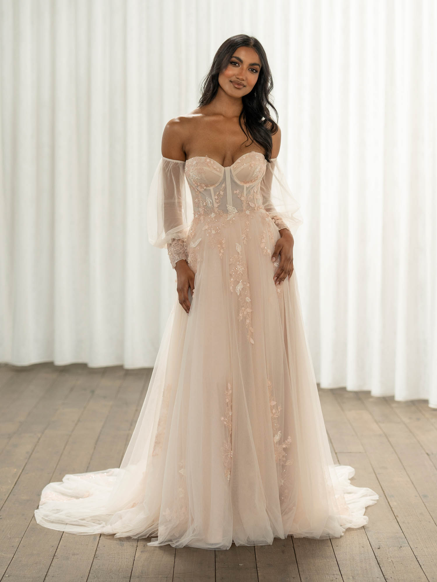 Woman wearing Turner by Madi Lane, a white wedding dress with sheer sleeves in a room with white curtains.