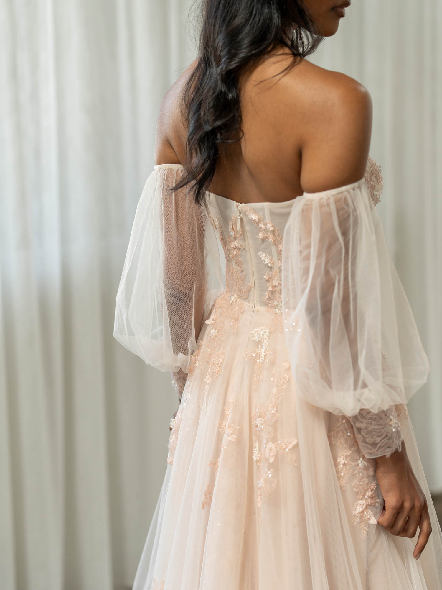 Woman wearing Turner by Madi Lane, in this photo shown in a blush pink off-shoulder dress with sheer sleeves against a white curtain background