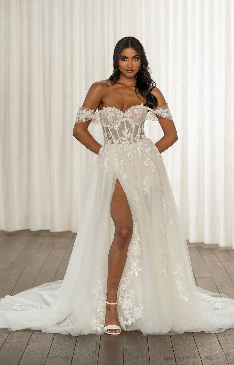 Woman wearing Twyla by Madi Lane, a white off-shoulder wedding dress with a high slit on a wooden floor.