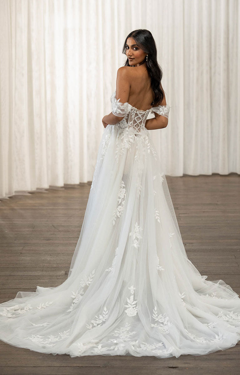 Woman wearing Twyla by Madi Lane, a white wedding dress with lace details in a room with white curtains.