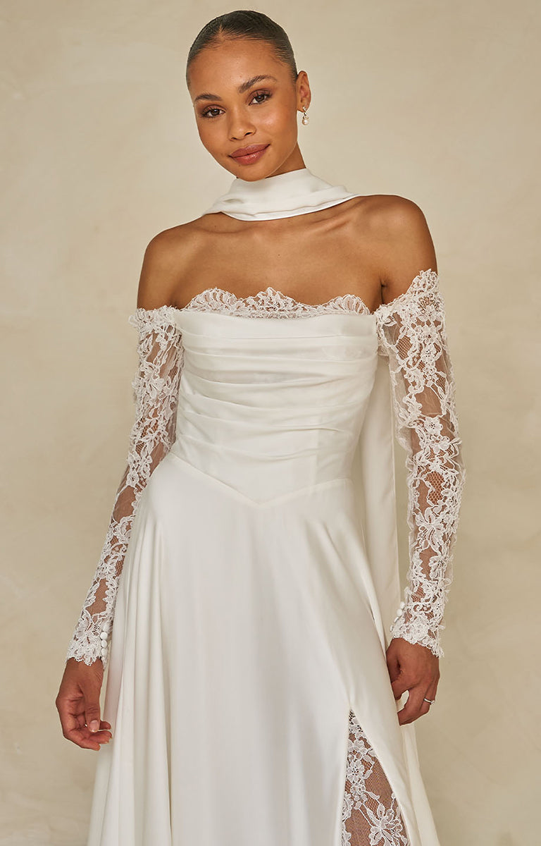 Woman wearing Ward by Madi Lane Bridal - a white lace wedding dress with long sleeves on a beige background