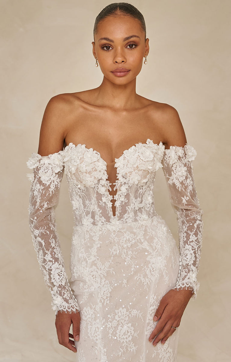 Woman wearing a white lace off-shoulder gown with long sleeves on a beige background
