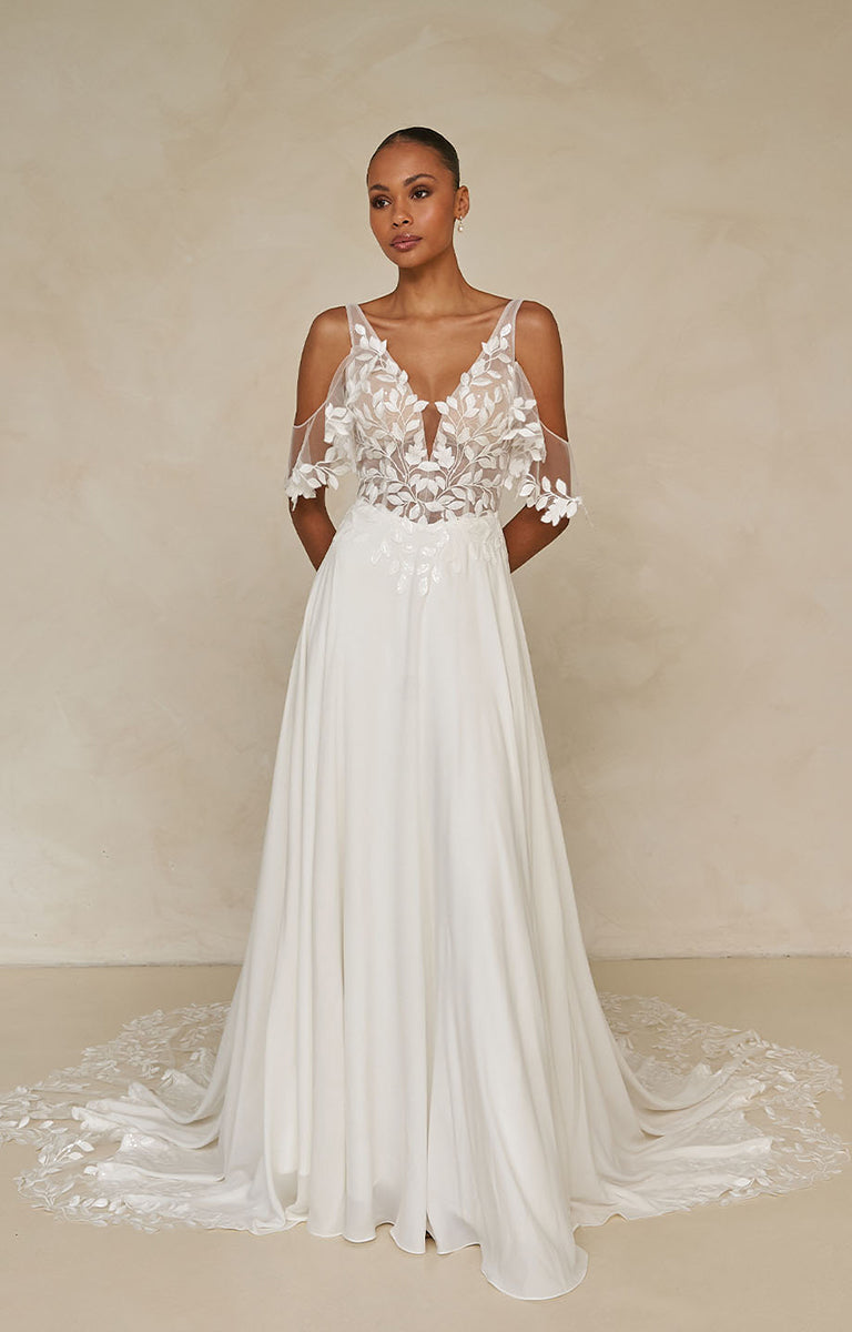 Woman wearing Wilmott by Madi Lane - a white wedding dress with lace detailing against a beige background