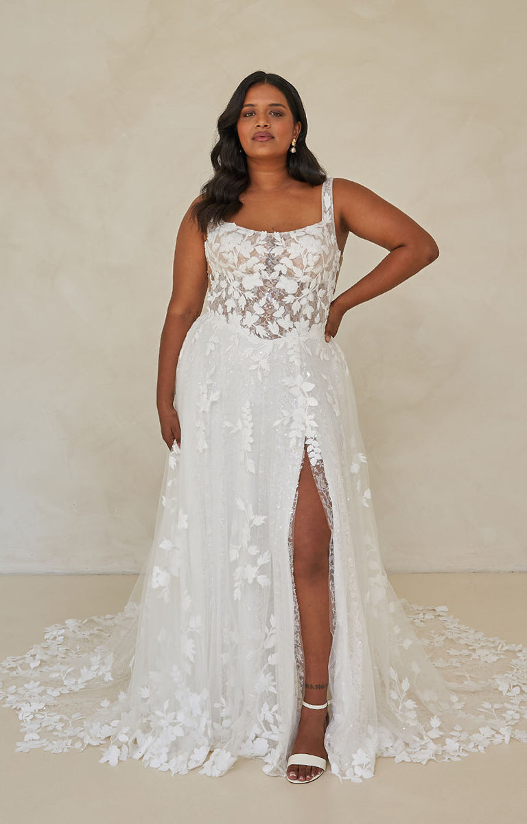 Woman wearing Wimmer by Madi Lane, a white lace wedding dress with a slit on a plain background