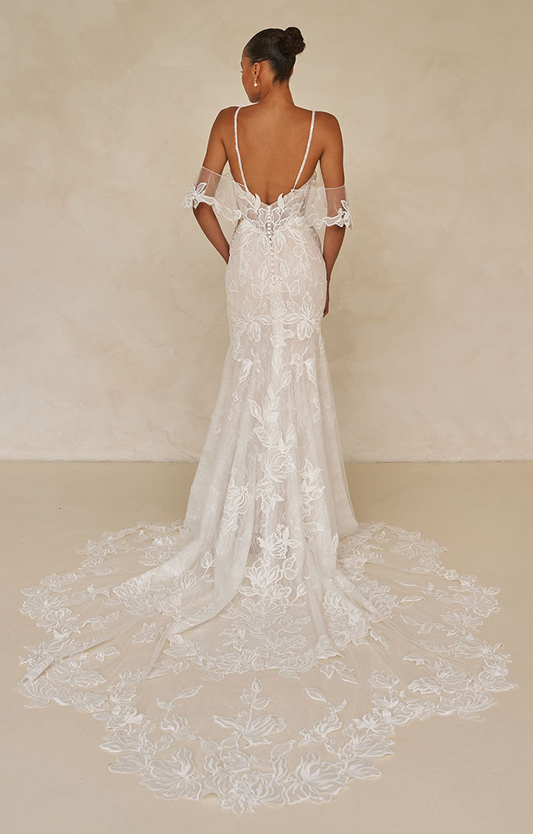 Woman wearing Winona by Madi Lane a White lace wedding dress with a long train on a plain background
