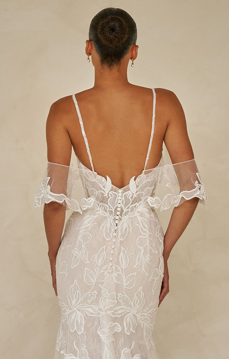 Back view of a woman wearing Winona by Madi Lane, a white lace wedding dress with sheer sleeves on a beige background