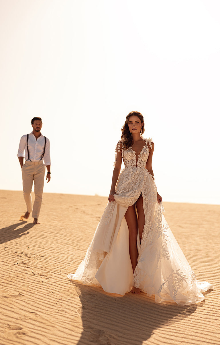 Woman in a white lace wedding dress standing in the desert with a man in formal attire behind her.