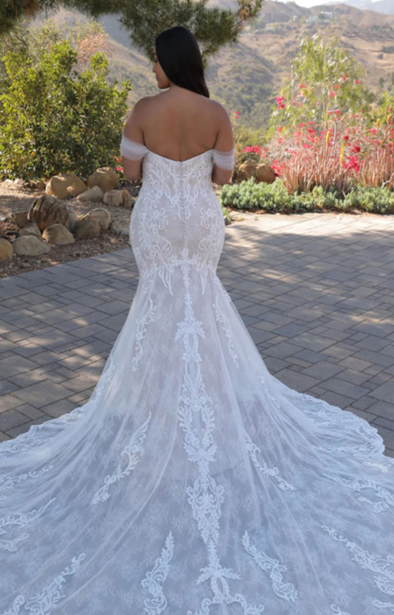 Woman in Athénais by ÉLYSÉE Edition - Floral Baroque Lace Wedding Dress with Sculpted Train and Detachable Sleeves standing outdoors with mountains and flowers in the background