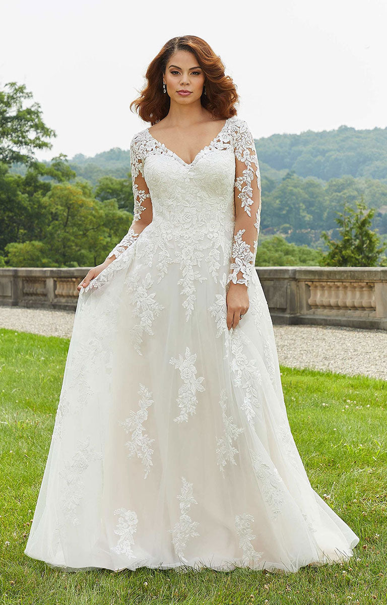 Woman wearing Emilia Wedding Dress by Morilee - Style 3346 – Floral Lace A-Line Gown with Illusion Sleeves standing outdoors with greenery in the background