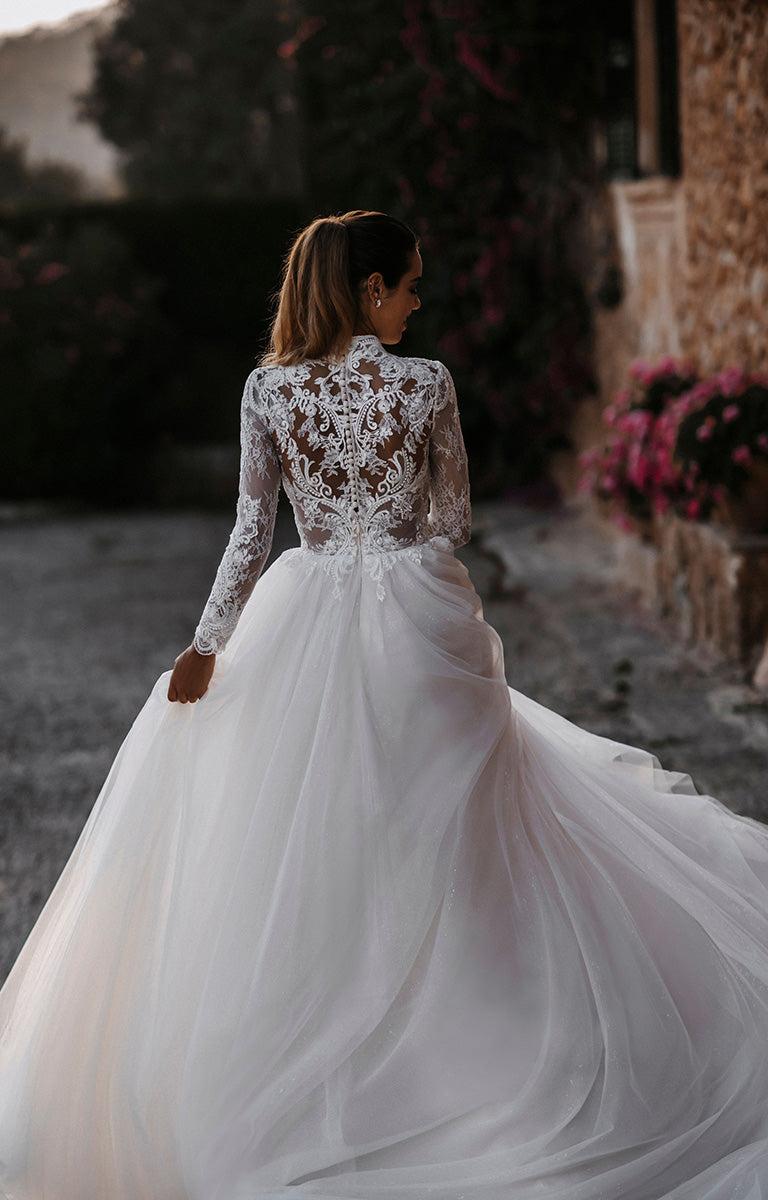 Woman in a white lace wedding dress standing outdoors with a stone building and flowers in the background.