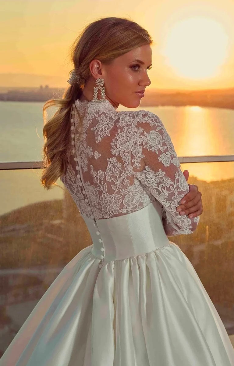 Woman in Legacy by Eddy K - Mikado Ballgown Wedding Dress with Lace Sleeves and Structured Waistband standing on a balcony with a sunset view.