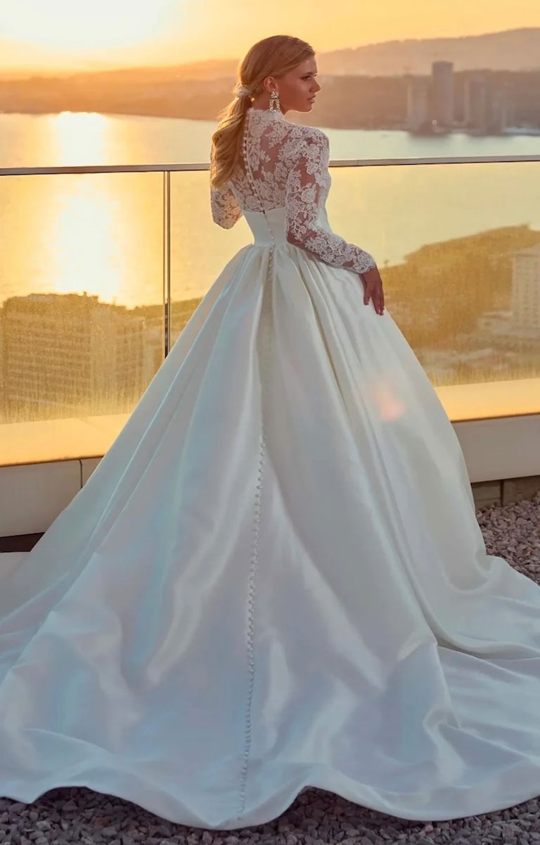 Woman in Legacy by Eddy K - Mikado Ballgown Wedding Dress with Lace Sleeves and Structured Waistband with lace sleeves standing on a balcony with a cityscape and sunset in the background.