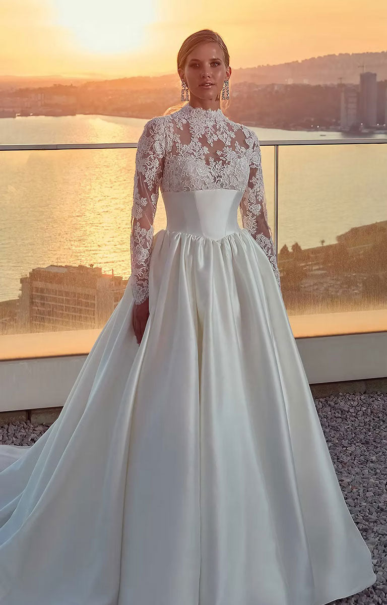 Woman in Legacy by Eddy K - Mikado Ballgown Wedding Dress with Lace Sleeves and Structured Waistband standing on a balcony with a sunset view.