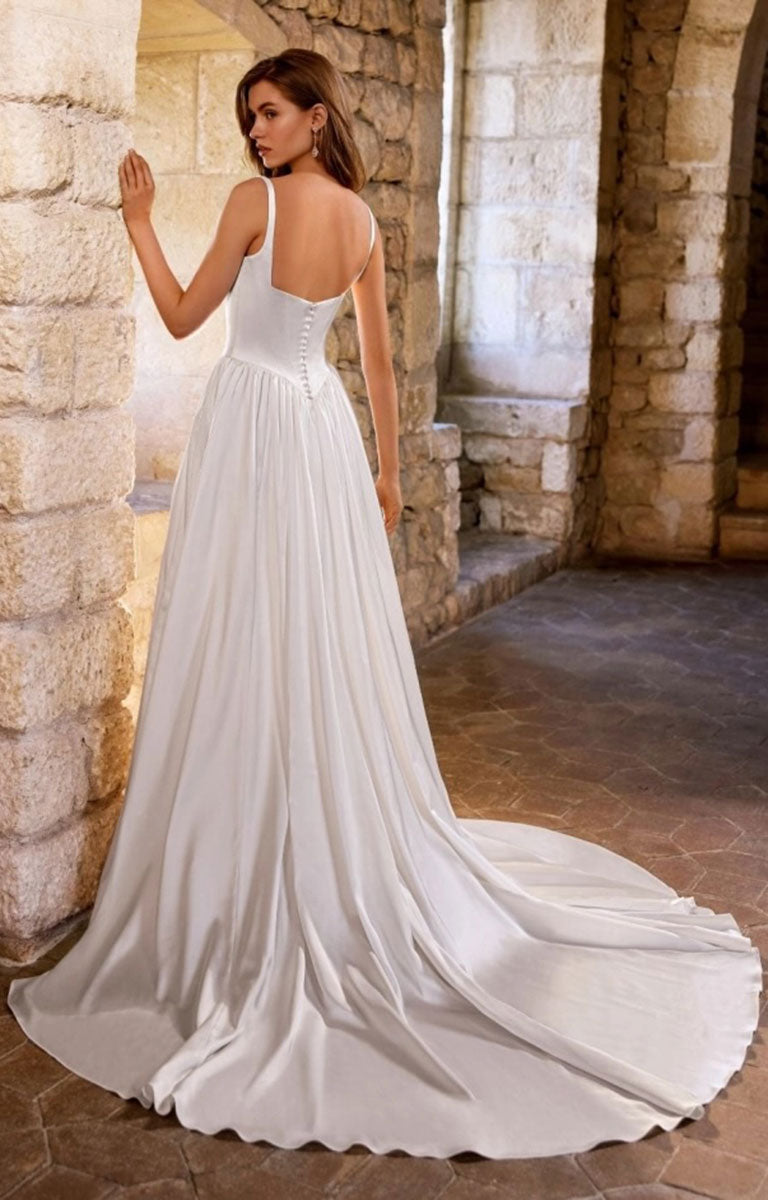 A woman wearing Gabbes by Étoile Bridal - Matte Satin A-Line Wedding Dress with Square Neckline and Basque Waist with a fitted corset-style bodice and a long flowing train, photographed in a stone archway corridor.