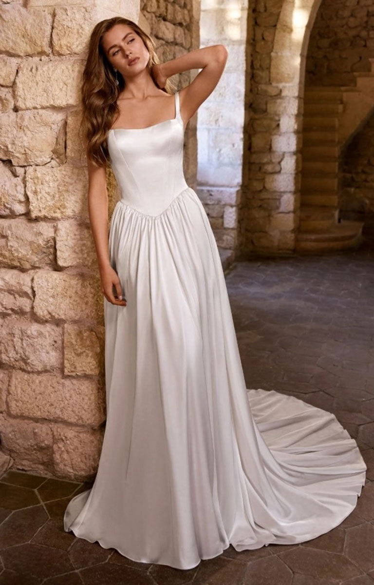 A woman modeling Gabbes by Étoile Bridal - Matte Satin A-Line Wedding Dress with Square Neckline and Basque Waist with thin straps, a fitted bodice, and a flowing skirt with a train, posed in front of a rustic stone wall and arched corridor.