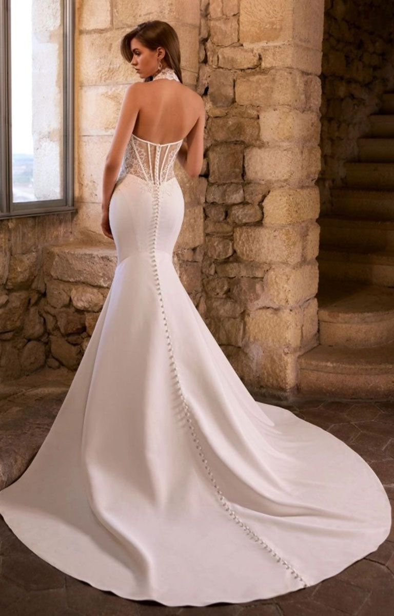 A woman wearing Gabrianna by Étoile Bridal - Fit-and-Flare Wedding Dress with Halter Neckline and Beaded Lace with an open back, lace-up corset-style waist, and long train, standing in a stone interior.