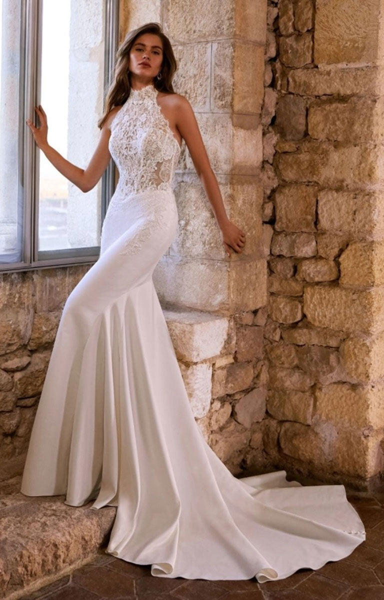 A woman models Gabrianna by Étoile Bridal - Fit-and-Flare Wedding Dress with Halter Neckline and Beaded Lace with a lace bodice, high neckline, and long train, posed against a rustic stone wall and window.