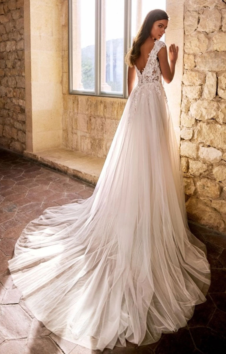 A woman wearing Gemmah by Étoile Bridal - Beaded Lace A-Line Wedding Dress with V-Neckline and Royal Train stands indoors beside a stone wall and window, showing the back of a white wedding dress with lace detailing and a long flowing train.